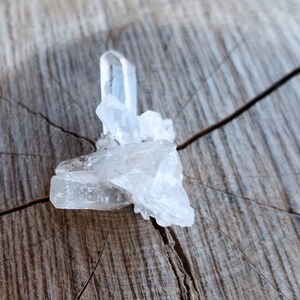 May include: A cluster of clear quartz crystals on a rough wooden surface. The crystals are arranged in a loose, organic shape, with some of the points facing upwards.