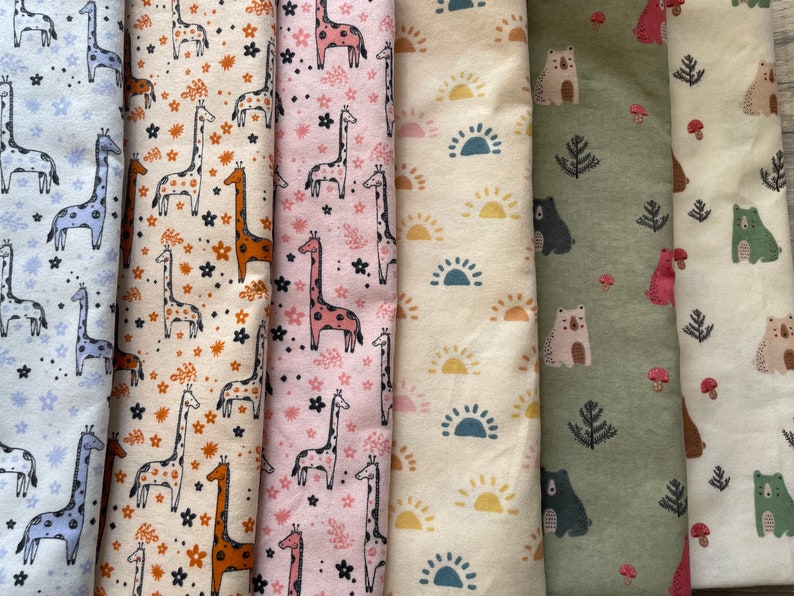 May include: A collection of six fabric swatches featuring different animal and nature-themed patterns. The swatches include a light blue fabric with giraffes, a beige fabric with giraffes, a pink fabric with giraffes, a beige fabric with suns, a green fabric with bears and trees, and a beige fabric with bears and mushrooms.