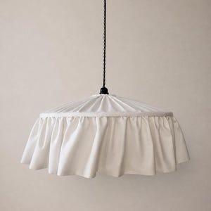 Op de afbeelding: Een witte stoffen hanglamp met een geplooid bovendeel en een brede, geplooide rand rond de basis. De lamp hangt aan een dun zwart snoer en ketting, met een zwart armatuur aan de bovenkant waar de stof het snoer ontmoet.