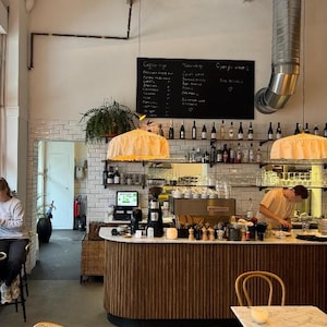 May include: A cafe interior featuring a curved wooden bar with a white marble countertop, a black menu board with white text, two ruffled pale yellow pendant lights, and shelves stocked with various glass bottles and glassware against a white brick wall.