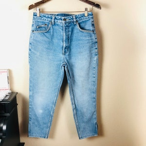 May include: Light blue denim jeans with a high waist and a straight leg. The jeans have front and back pockets, a button and zipper closure, and a small slit at the ankle. The jeans are hanging on a wooden hanger.