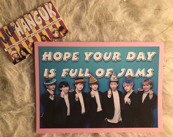 May include: A greeting card with a blue background and pink border. The card features a group of seven men wearing party hats and holding party favours. The text on the card reads "HOPE YOUR DAY IS FULL OF JAMS".