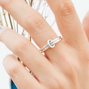 Può includere: Un anello color argento con un design a nodo. L'anello è su un dito con smalto grigio chiaro. Lo sfondo è bianco, con un vaso blu e fiori viola.