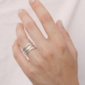 Può includere: Un anello d'argento con un design ondulato e organico. L'anello è indossato sul dito medio di una mano.