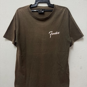 K&ouml;nnte beinhalten: Braunes T-Shirt mit dem Fender-Logo in wei&szlig;er Schrift auf der Vorderseite. Das T-Shirt h&auml;ngt an einem schwarzen Kleiderb&uuml;gel. Das T-Shirt hat kurze &Auml;rmel und einen Rundhalsausschnitt.