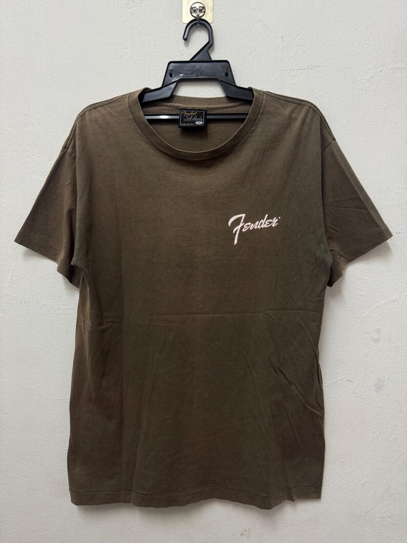 K&ouml;nnte beinhalten: Braunes T-Shirt mit dem Fender-Logo in wei&szlig;er Schrift auf der Vorderseite. Das T-Shirt h&auml;ngt an einem schwarzen Kleiderb&uuml;gel. Das T-Shirt hat kurze &Auml;rmel und einen Rundhalsausschnitt.