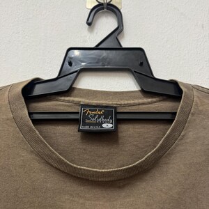 K&ouml;nnte beinhalten: Ein braunes Fender Solidbody T-Shirt, Gr&ouml;&szlig;e L, h&auml;ngt an einem schwarzen Kleiderb&uuml;gel. Auf dem Etikett des Shirts steht "Fender Solidbody" und "Made in USA". Das Shirt ist einfarbig und hat einen Rundhalsausschnitt.