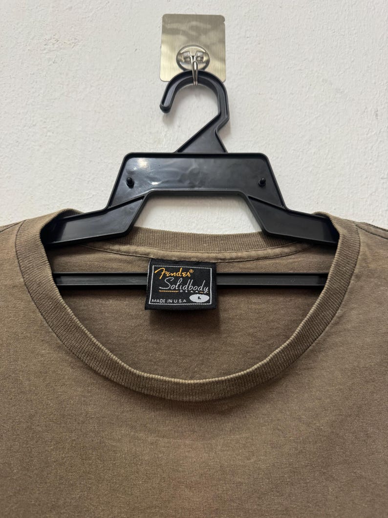 K&ouml;nnte beinhalten: Ein braunes Fender Solidbody T-Shirt, Gr&ouml;&szlig;e L, h&auml;ngt an einem schwarzen Kleiderb&uuml;gel. Auf dem Etikett des Shirts steht "Fender Solidbody" und "Made in USA". Das Shirt ist einfarbig und hat einen Rundhalsausschnitt.