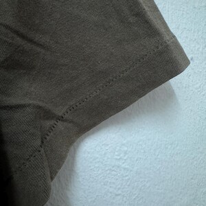 K&ouml;nnte beinhalten: Nahaufnahme eines Abschnitts eines braunen T-Shirts. Der Stoff ist strukturiert und zeigt den Saum mit einer sichtbaren Naht. Das Shirt besteht aus einem weichen Material und ist wahrscheinlich f&uuml;r den Freizeitgebrauch.