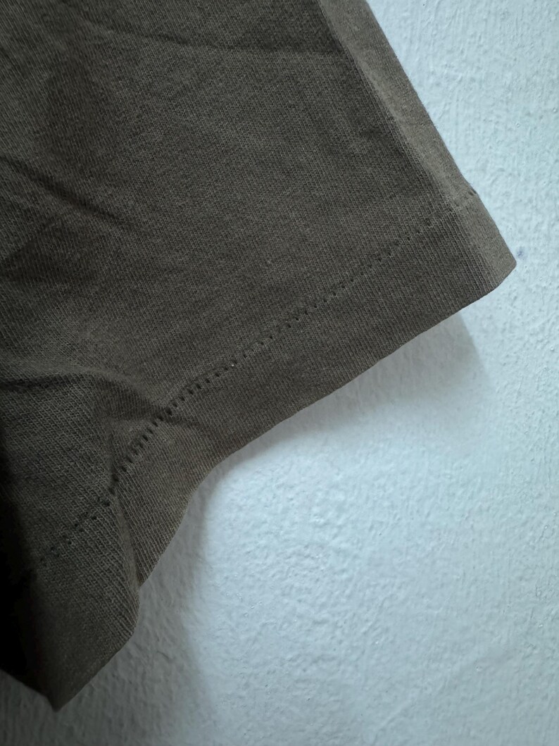K&ouml;nnte beinhalten: Nahaufnahme eines Abschnitts eines braunen T-Shirts. Der Stoff ist strukturiert und zeigt den Saum mit einer sichtbaren Naht. Das Shirt besteht aus einem weichen Material und ist wahrscheinlich f&uuml;r den Freizeitgebrauch.