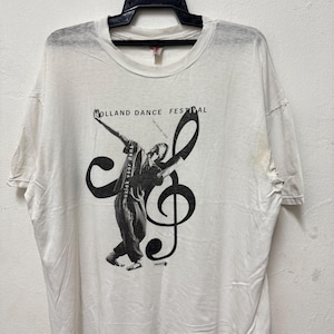 Puede incluir: Camiseta blanca con un gráfico en blanco y negro de un bailarín y una clave de sol. El texto "HOLLAND DANCE FESTIVAL" y "muziek voor dans" están impresos en la camiseta. La camiseta está colgada en una percha negra.