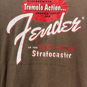 K&ouml;nnte beinhalten: Braunes T-Shirt mit einem Fender-Logo in Wei&szlig; und Rot. Der Text enth&auml;lt "Synchronized Tremolo Action..." und "Stratocaster" in einem Vintage-Stil. Das Shirt hat einen klassischen, abgenutzten Look.