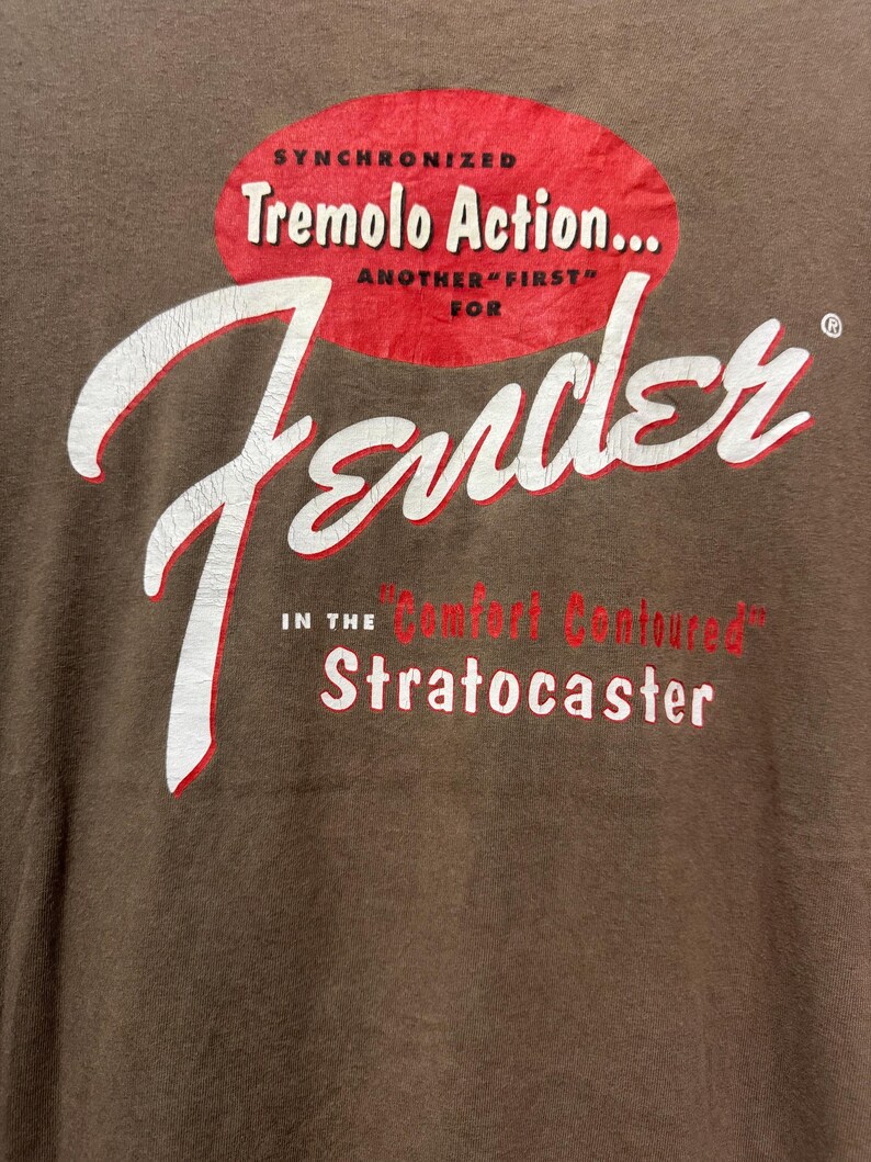 K&ouml;nnte beinhalten: Braunes T-Shirt mit einem Fender-Logo in Wei&szlig; und Rot. Der Text enth&auml;lt "Synchronized Tremolo Action..." und "Stratocaster" in einem Vintage-Stil. Das Shirt hat einen klassischen, abgenutzten Look.