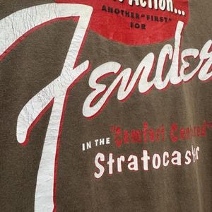 K&ouml;nnte beinhalten: Nahaufnahme eines braunen T-Shirts mit dem Fender-Logo in Wei&szlig; und Rot. Der Text enth&auml;lt "Tremolo Action" und "Stratocaster". Der Used-Look verleiht einen Vintage-Look.
