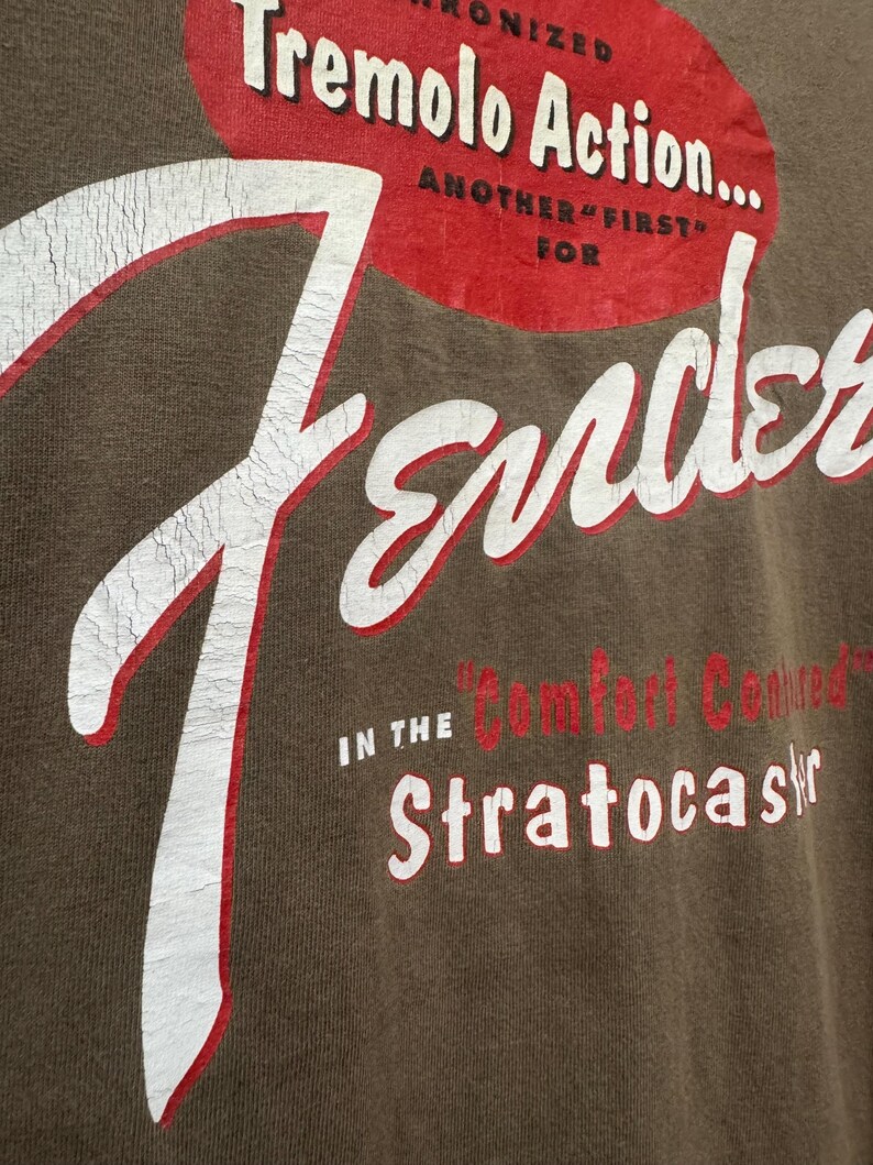 K&ouml;nnte beinhalten: Nahaufnahme eines braunen T-Shirts mit dem Fender-Logo in Wei&szlig; und Rot. Der Text enth&auml;lt "Tremolo Action" und "Stratocaster". Der Used-Look verleiht einen Vintage-Look.