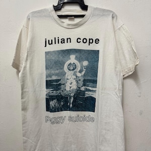 Può includere: Maglietta bianca con una stampa grafica in nero e blu di una persona con un cappello che tiene una sfera. Il testo "julian cope" e "peggy suicide" è stampato in nero sulla maglietta.