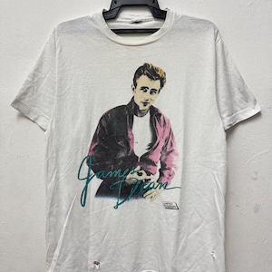 Puede incluir: Camiseta blanca con un gráfico de James Dean con una chaqueta rosa. El texto "James Dean" está escrito en guión turquesa. La camiseta tiene un aspecto vintage con algo de desgaste, incluyendo pequeños agujeros en la parte inferior.