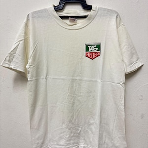 以下が含まれることがあります： クルーネックと半袖のオフホワイトのTシャツ。胸元には「TAG Heuer」の文字が入った緑と赤の小さなロゴが付いています。Tシャツは黒いハンガーにかかっています。