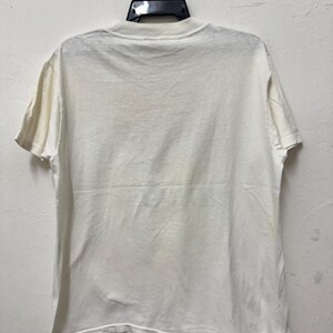 May include: A plain, off-white t-shirt hanging on a black hanger. The short-sleeved shirt is made of a soft fabric and has a crew neck. The back of the shirt is visible, showing the simple design.
