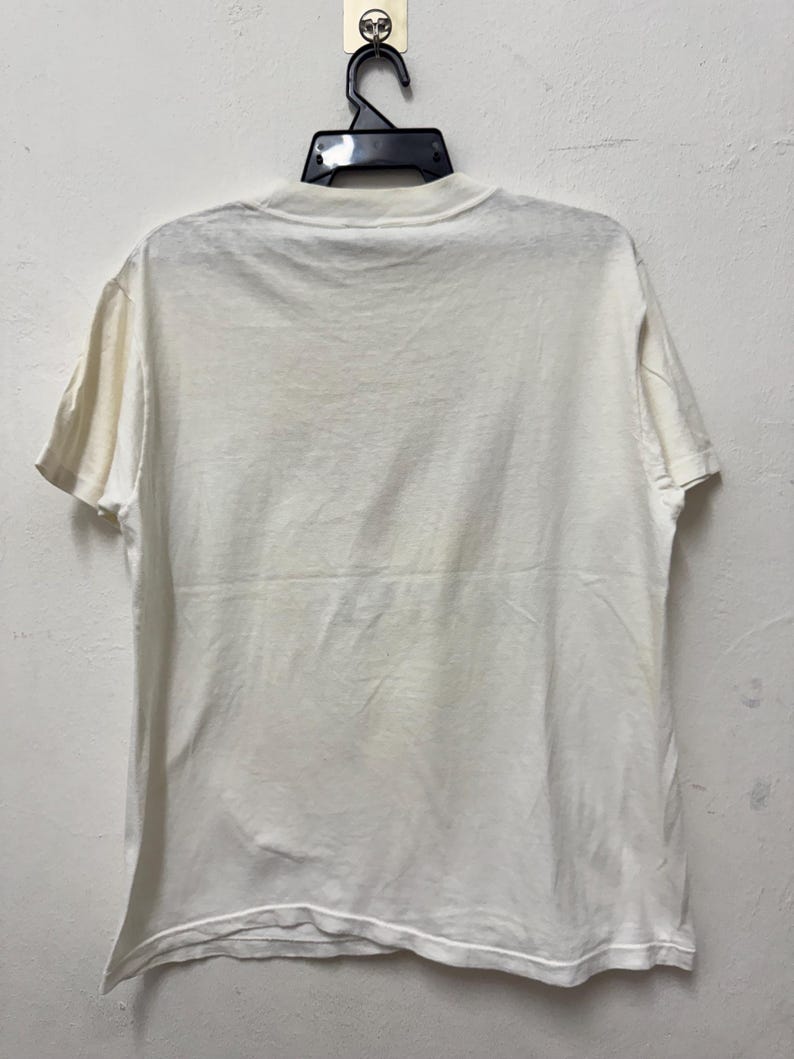 May include: A plain, off-white t-shirt hanging on a black hanger. The short-sleeved shirt is made of a soft fabric and has a crew neck. The back of the shirt is visible, showing the simple design.