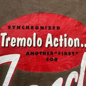 K&ouml;nnte beinhalten: Nahaufnahme eines braunen T-Shirts mit einem roten ovalen Grafikdesign. Die Grafik zeigt die Worte "SYNCHRONIZED Tremolo Action... ANOTHER "FIRST" FOR" in wei&szlig;em und schwarzem Text. Das T-Shirt hat einen Vintage-Look.