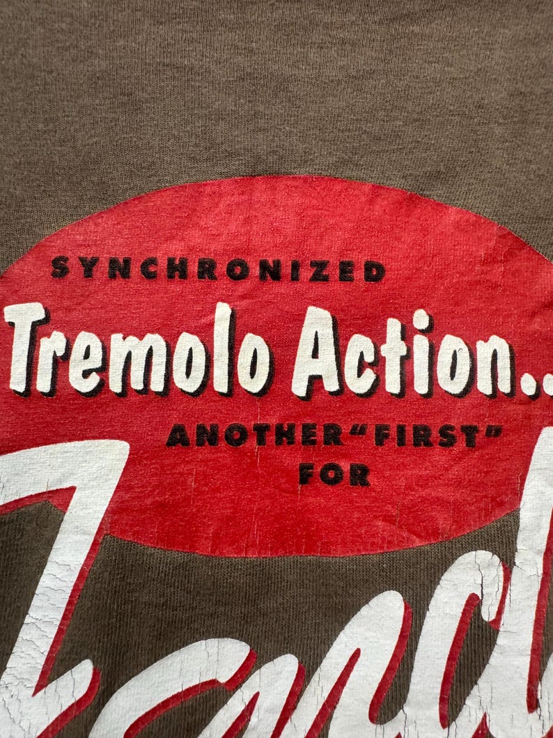 K&ouml;nnte beinhalten: Nahaufnahme eines braunen T-Shirts mit einem roten ovalen Grafikdesign. Die Grafik zeigt die Worte "SYNCHRONIZED Tremolo Action... ANOTHER "FIRST" FOR" in wei&szlig;em und schwarzem Text. Das T-Shirt hat einen Vintage-Look.
