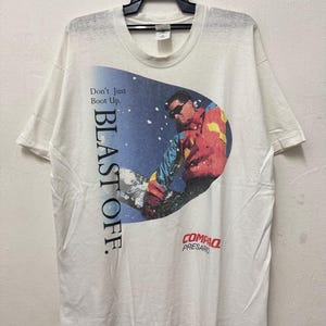Peut inclure: T-shirt blanc avec un graphisme d'un snowboarder portant des lunettes de soleil et une veste colorée. Le t-shirt affiche le texte "Don't Just Boot Up. BLAST OFF." et le logo Compaq Presario.