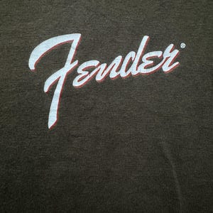 K&ouml;nnte beinhalten: Dunkelbraunes T-Shirt mit dem Wort "Fender" in wei&szlig;er Schrift mit roter Umrandung. Das Fender-Logo ist ein klassisches Design, wahrscheinlich f&uuml;r Musikliebhaber.