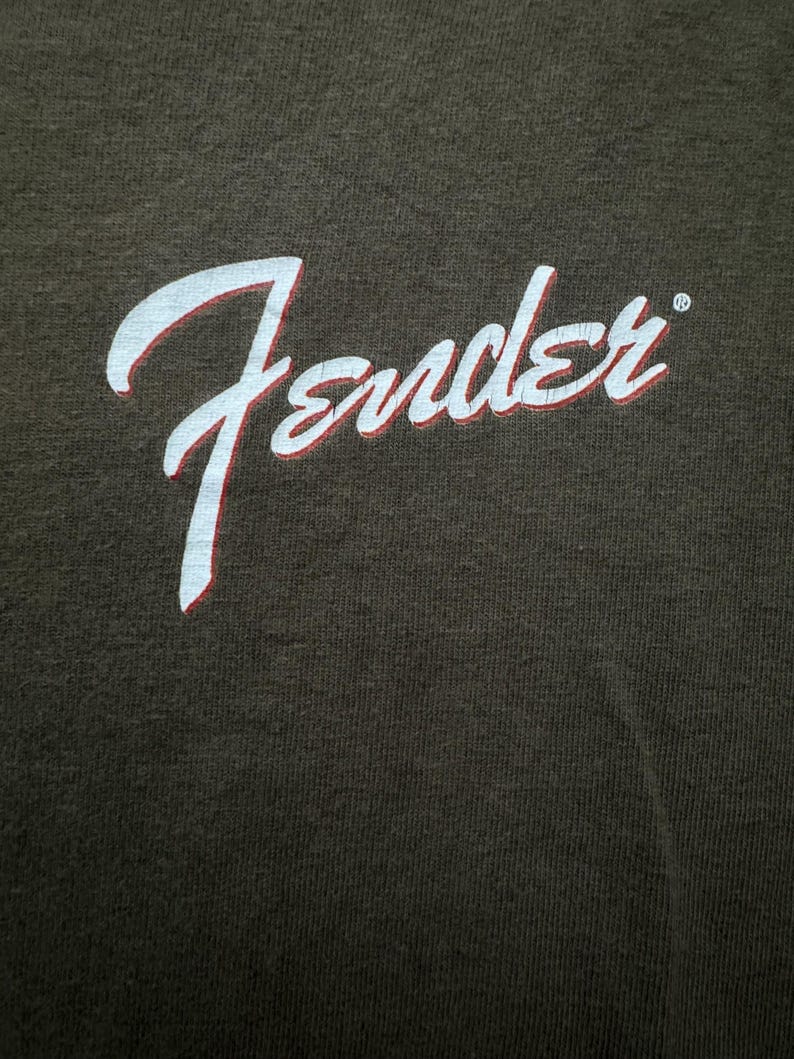 K&ouml;nnte beinhalten: Dunkelbraunes T-Shirt mit dem Wort "Fender" in wei&szlig;er Schrift mit roter Umrandung. Das Fender-Logo ist ein klassisches Design, wahrscheinlich f&uuml;r Musikliebhaber.