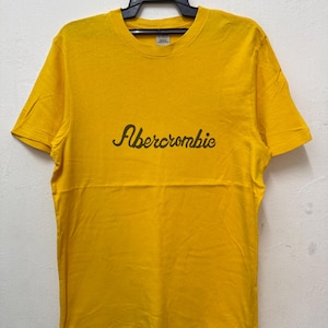 May include: A bright yellow short-sleeved t-shirt featuring the word Abercrombie printed in a dark brown cursive script across the centre of the chest. The garment has a crew neckline and is displayed on a black plastic hanger against a white wall.