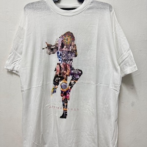 Op de afbeelding: Wit t-shirt met een collage-ontwerp in de vorm van een persoon. De collage bestaat uit verschillende afbeeldingen en kleuren. De tekst "JETHRO TULL" is rood onderaan afgedrukt. Het shirt hangt aan een zwarte hanger.