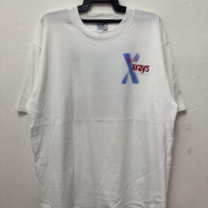 May include: White t-shirt with the text "Ray-Ban Xrays" in blue and red. The shirt is hanging on a black hanger. The t-shirt is a Gildan brand.