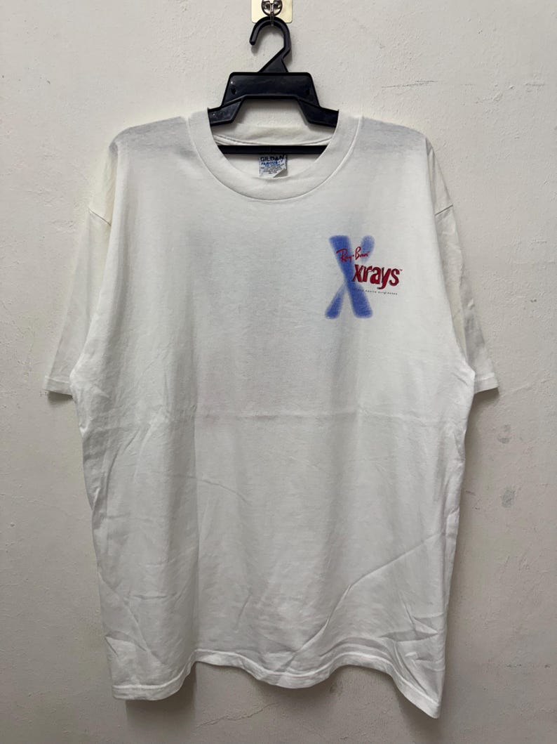 May include: White t-shirt with the text "Ray-Ban Xrays" in blue and red. The shirt is hanging on a black hanger. The t-shirt is a Gildan brand.
