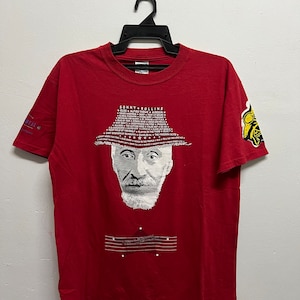 Puede incluir: Una camiseta roja con un retrato en blanco y negro de Sonny Rollins. La camiseta muestra el texto "SONNY ROLLINS" encima del retrato y una lista de títulos de canciones en forma de sombrero. La manga tiene un gráfico amarillo y negro.