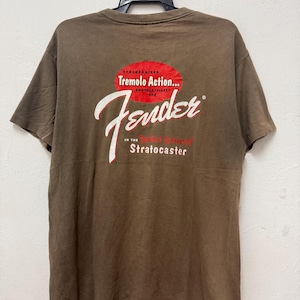 K&ouml;nnte beinhalten: Braunes T-Shirt mit dem Fender-Logo in wei&szlig;er Schrift und dem Text "Synchronized Tremolo Action... Another First for Stratocaster" in Rot und Wei&szlig;. Das T-Shirt h&auml;ngt an einem schwarzen Kleiderb&uuml;gel.
