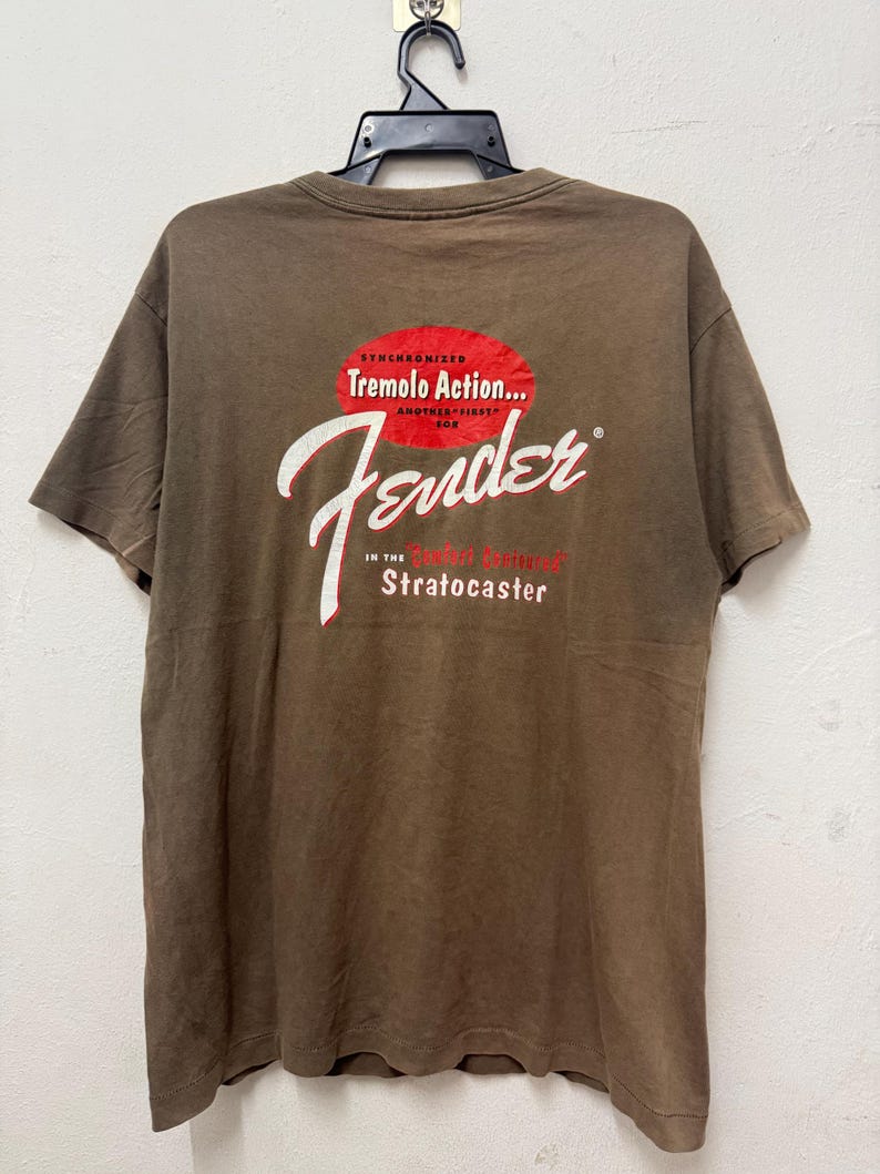 K&ouml;nnte beinhalten: Braunes T-Shirt mit dem Fender-Logo in wei&szlig;er Schrift und dem Text "Synchronized Tremolo Action... Another First for Stratocaster" in Rot und Wei&szlig;. Das T-Shirt h&auml;ngt an einem schwarzen Kleiderb&uuml;gel.
