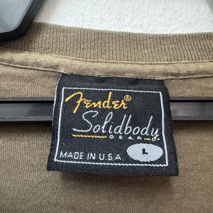 K&ouml;nnte beinhalten: Nahaufnahme eines braunen T-Shirt-Kragens mit einem schwarzen Etikett. Auf dem Etikett steht "Fender Solidbody Gear" in goldener und wei&szlig;er Schrift, darunter "MADE IN U.S.A.". Die Gr&ouml;&szlig;e "L" ist ebenfalls sichtbar.