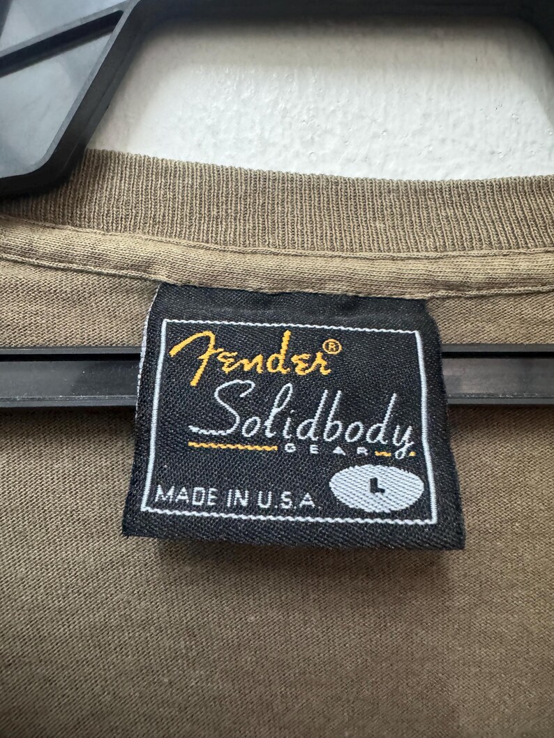 K&ouml;nnte beinhalten: Nahaufnahme eines braunen T-Shirt-Kragens mit einem schwarzen Etikett. Auf dem Etikett steht "Fender Solidbody Gear" in goldener und wei&szlig;er Schrift, darunter "MADE IN U.S.A.". Die Gr&ouml;&szlig;e "L" ist ebenfalls sichtbar.