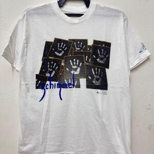 Puede incluir: Camiseta blanca con un diseño gráfico de múltiples huellas de manos en blanco y negro sobre un fondo oscuro. La palabra "Schimmel" está impresa en azul. La camiseta está colgada de una percha negra.