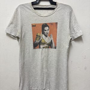May include: A light gray t-shirt featuring a graphic of a woman in a yellow top with her hands up. The graphic is set against an orange background. The word "AMY" is printed in the upper left corner of the graphic.