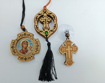 May include: Three wooden car charms with religious imagery. The first charm is a cross with the inscription "IC XC NIKA" and a suction cup for hanging. The second charm is a cross with a depiction of the Virgin Mary and baby Jesus. The third charm is a cross with a green gem and a black tassel.
