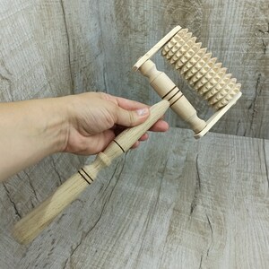 May include: A wooden massage roller with a handle and a spiked wheel. The roller is made of light brown wood and has a dark brown handle. The wheel is covered in small, sharp spikes.