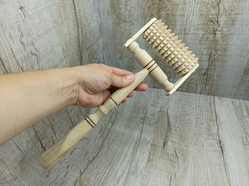 May include: A wooden massage roller with a handle and a spiked wheel. The roller is made of light brown wood and has a dark brown handle. The wheel is covered in small, sharp spikes.