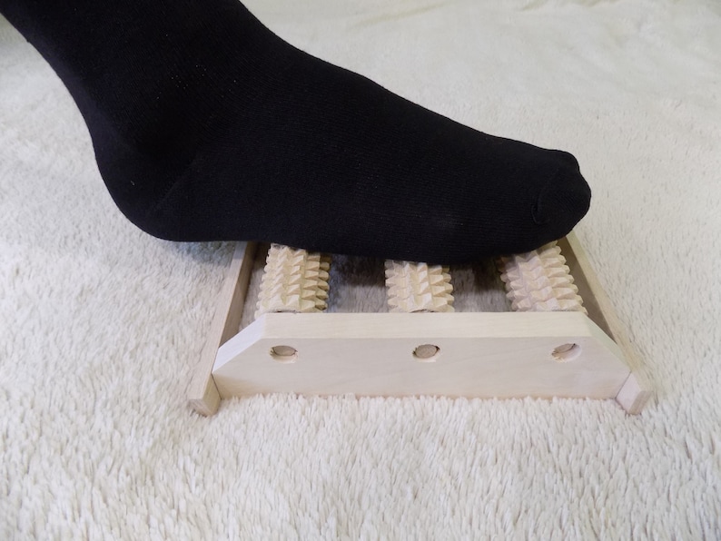 May include: A wooden foot roller with three rows of rounded spikes. A foot wearing a black sock is rolling over the spikes.