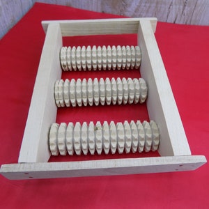 May include: A wooden foot roller with three rows of spiked rollers. The roller is made of natural wood and has a light brown colour.