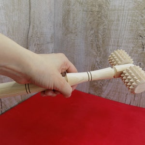 May include: A wooden rolling pin with two spiked heads. The rolling pin is held in a hand and is resting on a red surface.