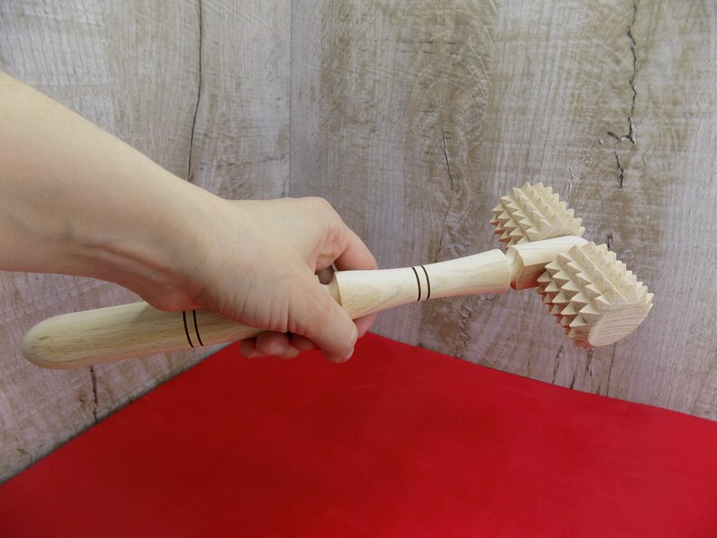 May include: A wooden rolling pin with two spiked heads. The rolling pin is held in a hand and is resting on a red surface.