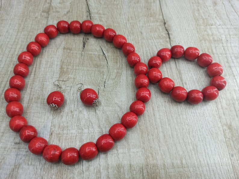 May include: A red wooden bead necklace, bracelet, and earrings set. The necklace and bracelet are made of large, round beads. The earrings are simple silver hoops with a red bead at the end.