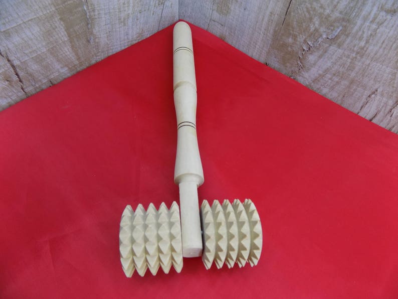 May include: A wooden rolling pin with two textured wheels. The wheels have a starburst pattern and are made of light brown wood. The rolling pin is on a red surface.