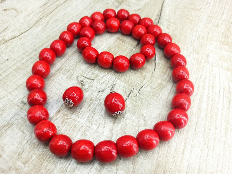 May include: A red wooden bead necklace, bracelet, and earrings set. The necklace and bracelet are made of large, round beads. The earrings are simple and dangle.